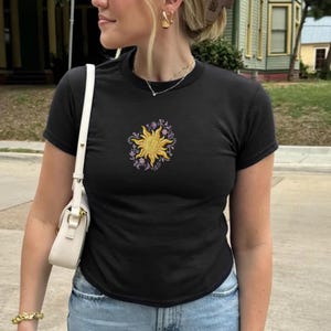 May include: Black crew neck t-shirt with a gold sun and purple floral embroidery on the front. The shirt is short-sleeved and made of a soft material. The shirt is paired with blue jeans.