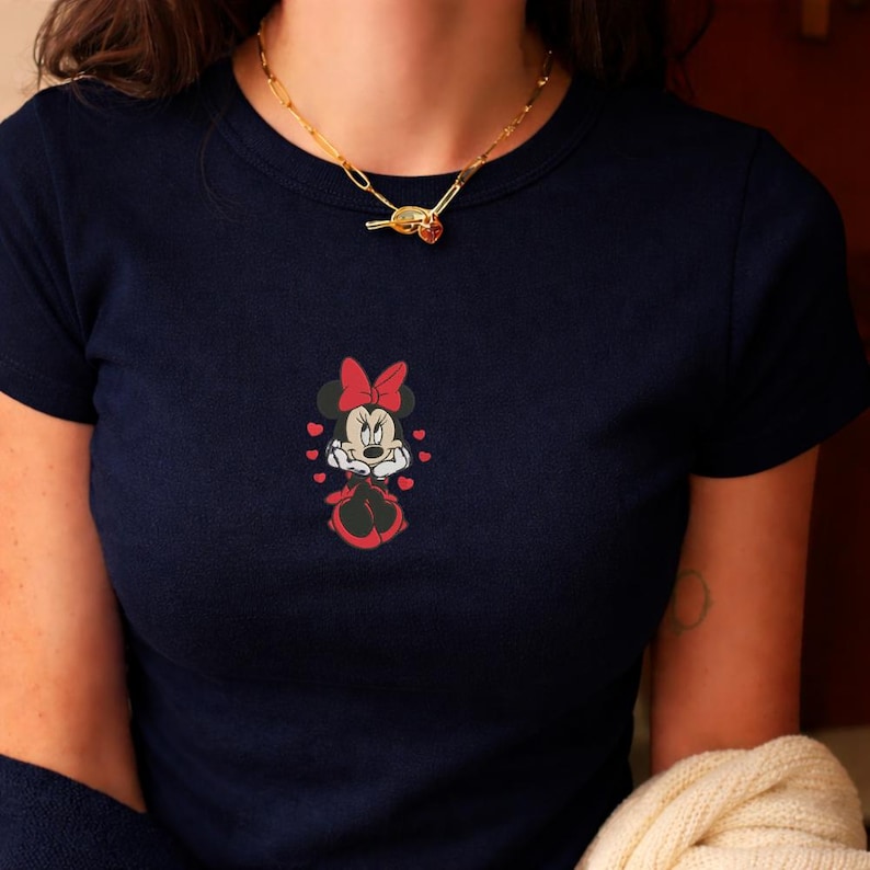 Peut inclure: T-shirt bleu marine avec un motif brod&eacute; de Minnie Mouse. Le motif comprend un n&oelig;ud rouge, des c&oelig;urs et le personnage de Minnie Mouse en noir et blanc. Le t-shirt a des manches courtes et un col rond.