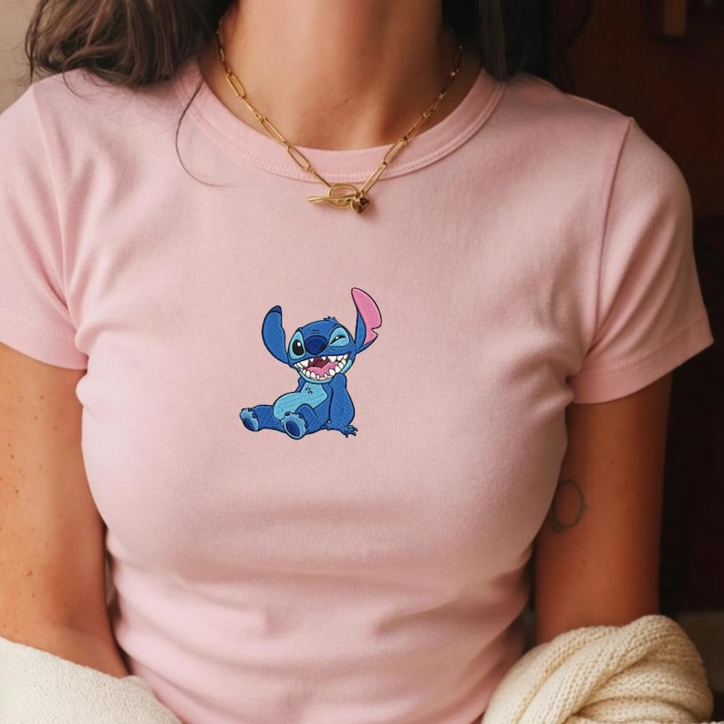 May include: A light pink short-sleeved t-shirt featuring a small embroidered graphic of the blue character Stitch from Lilo and Stitch sitting down and smiling. The shirt has a crew neckline and is paired with a gold-toned chain link necklace.