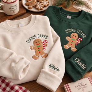 以下が含まれることがあります： ジンジャーブレッドマンと「COOKIE BAKER」の刺繍が入ったクリーム色のスウェットシャツと、ジンジャーブレッドマンと「COOKIE TESTER」の刺繍が入ったダークグリーンのスウェットシャツ。どちらも袖口に名前が刺繍されています。