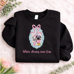 Puede incluir: Una sudadera negra de manga larga con un marco ovalado bordado en azul claro con un lazo rosa en la parte superior. Dentro del marco hay dos personajes de dibujos animados y un castillo. Debajo del dise&ntilde;o, el texto dice Where dreams come true.