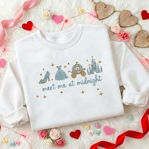 Puede incluir: Sudadera blanca con dise&ntilde;o bordado que presenta un tac&oacute;n alto, un vestido, un carruaje y un castillo. El texto "meet me at midnight" est&aacute; bordado en azul. Tambi&eacute;n hay cruces doradas bordadas.