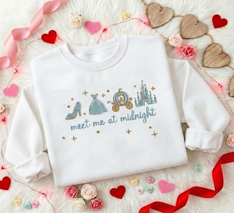 Puede incluir: Sudadera blanca con dise&ntilde;o bordado que presenta un tac&oacute;n alto, un vestido, un carruaje y un castillo. El texto "meet me at midnight" est&aacute; bordado en azul. Tambi&eacute;n hay cruces doradas bordadas.