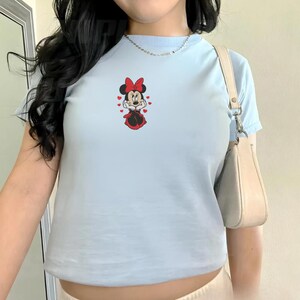 Peut inclure: T-shirt bleu clair orn&eacute; d'un motif Minnie Mouse. Le motif repr&eacute;sente Minnie Mouse dans une robe et un n&oelig;ud rouges, entour&eacute;e de c&oelig;urs rouges. Le t-shirt a des manches courtes et un col rond.