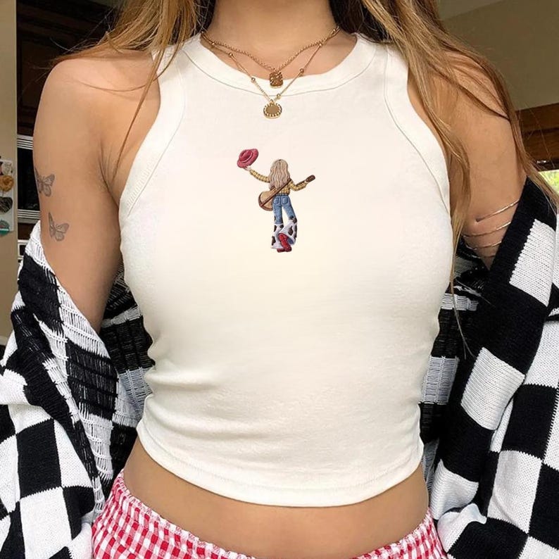 May include: A cream-colored sleeveless tank top featuring a small graphic print of a person with long blonde hair wearing a yellow shirt, blue jeans, and cow-print bell-bottoms, holding a guitar and a red hat, centered on the chest area.