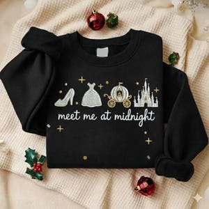 Puede incluir: Sudadera negra con dise&ntilde;o bordado que presenta un tac&oacute;n alto, un vestido, un carruaje y un castillo. El texto "meet me at midnight" est&aacute; debajo. La sudadera est&aacute; sobre una manta color crema con adornos navide&ntilde;os.