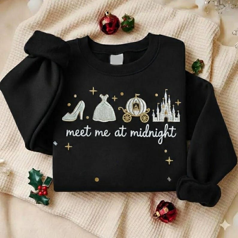 Puede incluir: Sudadera negra con dise&ntilde;o bordado que presenta un tac&oacute;n alto, un vestido, un carruaje y un castillo. El texto "meet me at midnight" est&aacute; debajo. La sudadera est&aacute; sobre una manta color crema con adornos navide&ntilde;os.