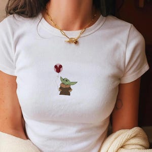 Puede incluir: Camiseta blanca con una imagen de dibujos animados de un personaje alien&iacute;gena verde sosteniendo un globo rosa. El alien&iacute;gena lleva una capa marr&oacute;n. Se lleva un collar de cadena dorada alrededor del cuello.
