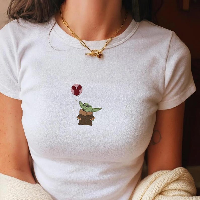 Puede incluir: Camiseta blanca con una imagen de dibujos animados de un personaje alien&iacute;gena verde sosteniendo un globo rosa. El alien&iacute;gena lleva una capa marr&oacute;n. Se lleva un collar de cadena dorada alrededor del cuello.