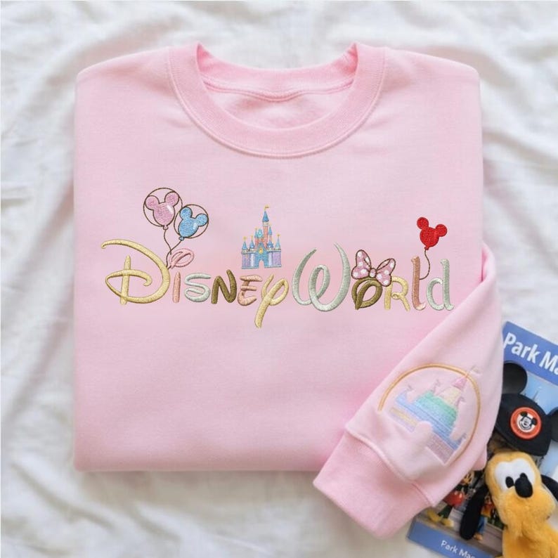 Puede incluir: Sudadera rosa claro con bordado "Disney World" y gr&aacute;ficos. El dise&ntilde;o incluye un castillo, globos y un lazo de Minnie Mouse. La manga presenta un dise&ntilde;o de castillo colorido.