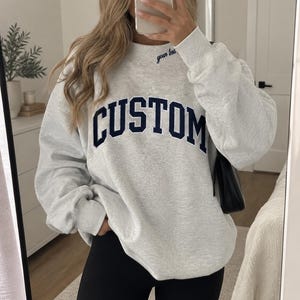 May include: A light grey crewneck sweatshirt featuring the word CUSTOM in large, dark navy blue block letters across the chest. The neckline includes the embroidered text your text in a smaller, cursive navy blue font on the collar area.
