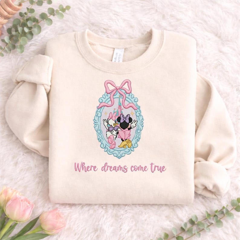Puede incluir: Una sudadera de cuello redondo color crema con un dise&ntilde;o bordado de Minnie Mouse y Daisy Duck dentro de un marco ovalado azul claro adornado con un lazo rosa. Debajo del gr&aacute;fico, el texto dice Where dreams come true en color rosa.
