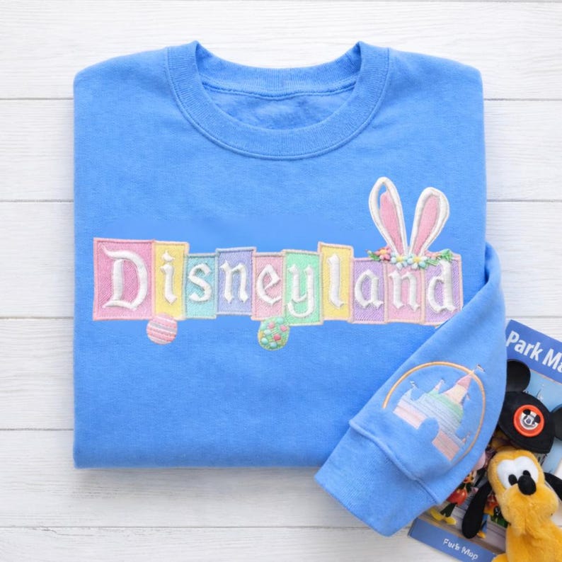 K&ouml;nnte beinhalten: Hellblaues Sweatshirt mit dem gestickten Wort "Disneyland" in Pastellfarben. Das Design umfasst Hasenohren und Ostereier. Der &Auml;rmel zeigt ein Schlossdesign. Eine Parkkarte und ein Mickey Mouse-Pl&uuml;schtier sind ebenfalls sichtbar.