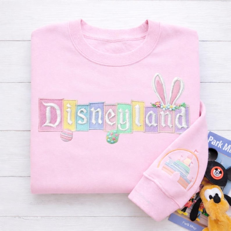K&ouml;nnte beinhalten: Ein hellrosa Sweatshirt mit dem Wort "Disneyland" in Pastellfarben gestickt. Das Design umfasst Hasenohren und Ostereier. Der &Auml;rmel zeigt ein Schlossdesign. Eine Parkkarte ist sichtbar.