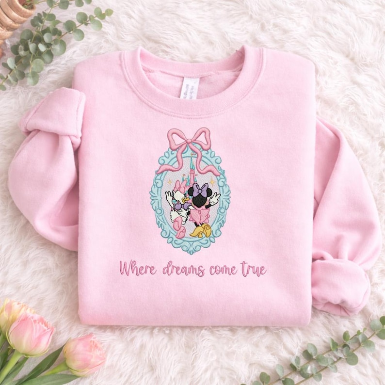 Puede incluir: Una sudadera de cuello redondo rosa claro con un dise&ntilde;o bordado de Minnie Mouse y Daisy Duck dentro de un marco ovalado azul claro adornado con un lazo rosa. Debajo del gr&aacute;fico, el texto dice Where dreams come true en color rosa.
