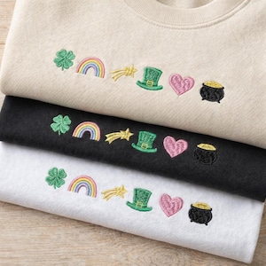 Puede incluir: Tres sudaderas plegadas en beige, negro y blanco, cada una bordada con diseños temáticos del Día de San Patricio. Los diseños incluyen un trébol verde, un arcoíris, una estrella fugaz, un sombrero verde, un corazón rosa y una olla de oro.