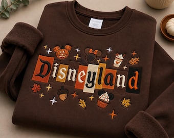 May include: A dark brown crewneck sweatshirt featuring embroidered autumn-themed icons and the word Disneyland in a stylized font. The design includes a pumpkin, mouse ears, a coffee cup, an acorn, a cupcake, and maple leaves with small stars.