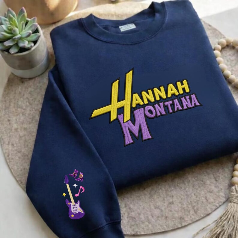 K&ouml;nnte beinhalten: Marineblaues Sweatshirt mit dem gestickten Text "Hannah Montana" in Gelb und Lila. Der linke &Auml;rmel zeigt eine lila Gitarre mit Noten. Das Sweatshirt ist aus weichem Material.
