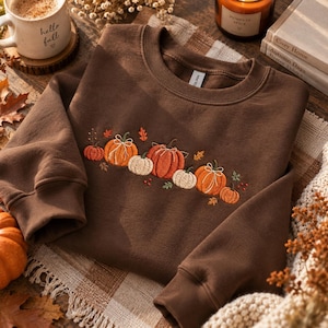 Puede incluir: Una sudadera de cuello redondo en color marrón oscuro con un diseño bordado en el pecho. El bordado muestra una disposición horizontal de varias calabazas en tonos naranja, crema y blanco, acentuadas con pequeñas hojas verdes y bayas rojas.