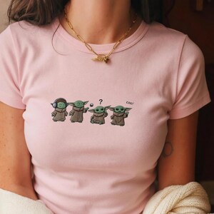 May include: A light pink t-shirt featuring a cartoon design of four Grogu characters in various poses. The design includes a Grogu with headphones, one holding a ball, one with a question mark, and one with the word "coo!"
