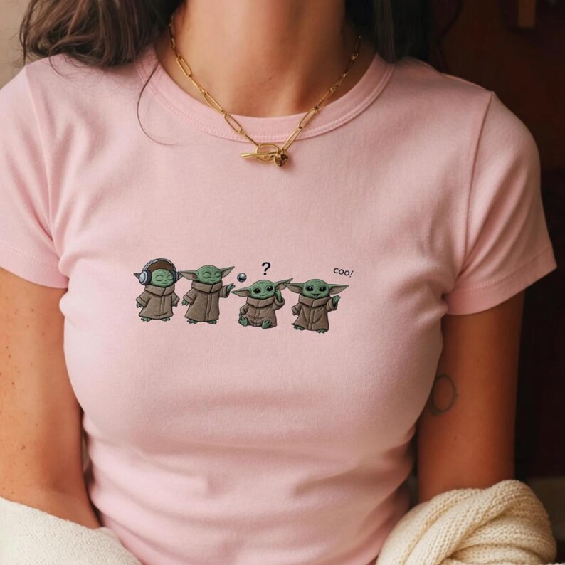 May include: A light pink t-shirt featuring a cartoon design of four Grogu characters in various poses. The design includes a Grogu with headphones, one holding a ball, one with a question mark, and one with the word "coo!"