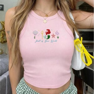 Puede incluir: Camiseta sin mangas rosa claro con cuello redondo. La parte superior presenta un dise&ntilde;o bordado de una concha, un tridente, una sirena, una estrella y coral, con el texto "Part of Your World" en cursiva.