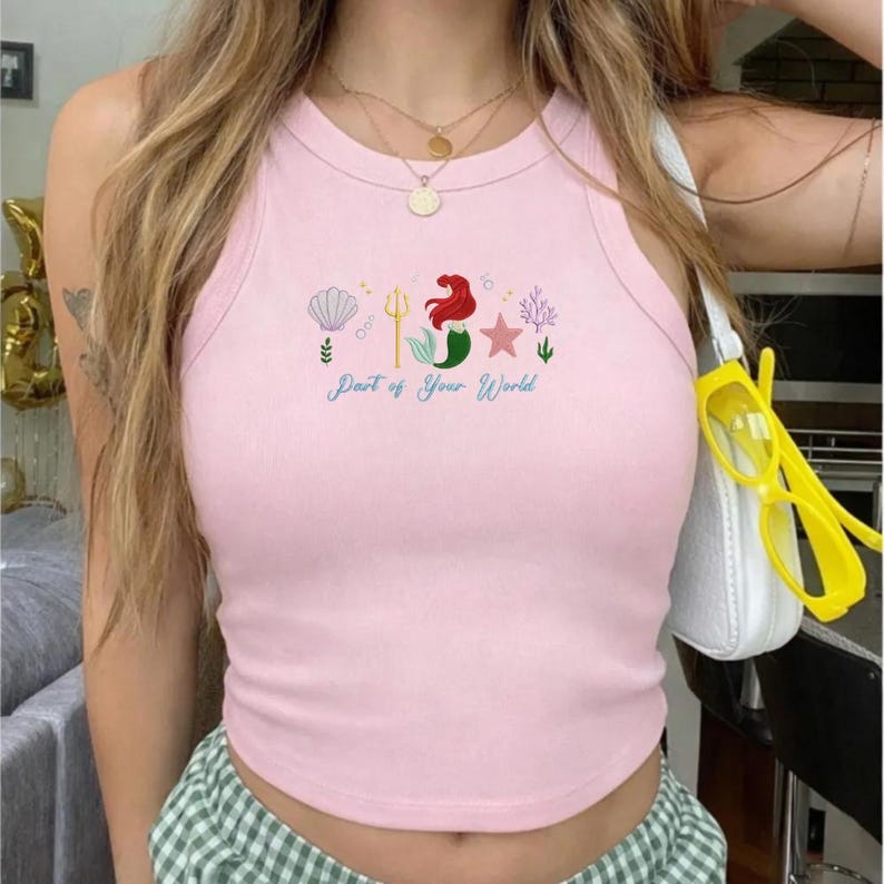 Puede incluir: Camiseta sin mangas rosa claro con cuello redondo. La parte superior presenta un dise&ntilde;o bordado de una concha, un tridente, una sirena, una estrella y coral, con el texto "Part of Your World" en cursiva.