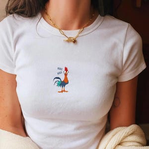 May include: White t-shirt with a colorful cartoon rooster graphic. The rooster has red, orange, blue, and green feathers and the text "HEI HEI" next to it. A gold chain necklace is also visible.