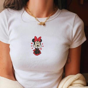 Peut inclure: T-shirt blanc avec un motif de Minnie Mouse. Le personnage de dessin anim&eacute; est repr&eacute;sent&eacute; dans une robe et un n&oelig;ud rouges, entour&eacute; de petits c&oelig;urs rouges. Le t-shirt a un col rond et des manches courtes.