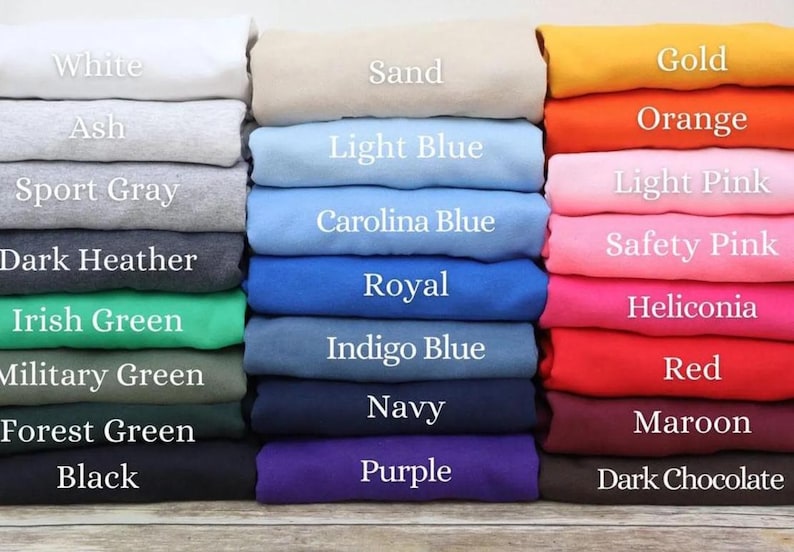 Puede incluir: Una colecci&oacute;n de prendas de tela dobladas apiladas en tres columnas, con una gran variedad de colores etiquetados con texto que incluye White, Sand, Gold, Ash, Orange, Light Blue, Light Pink, Dark Heather, Carolina Blue, Safety Pink, Irish Green, Royal, Heliconia, Military Green, Indigo Blue, Red, Forest Green, Navy, Maroon, Black, Purple y Dark Chocolate.