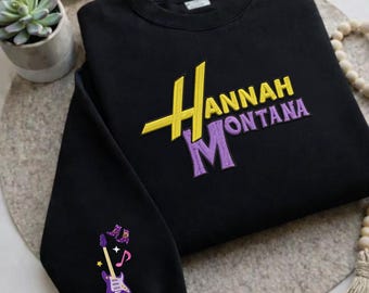 May include: Black sweatshirt with the text "Hannah Montana" in yellow and purple embroidery. The left sleeve features a purple guitar with musical notes and stars.