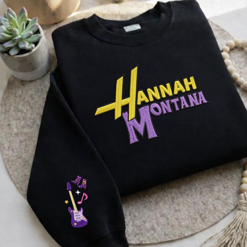 K&ouml;nnte beinhalten: Schwarzes Sweatshirt mit gesticktem Text "Hannah Montana" in Gelb und Lila. Der linke &Auml;rmel zeigt eine lila Gitarren-Grafik mit Noten und Sternen.