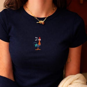 May include: Navy blue t-shirt featuring a colorful rooster graphic with the text "HEI HEI". The rooster has red, blue, and orange details. The shirt has short sleeves and a round neckline. A gold necklace is visible at the top.