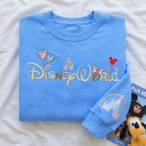 Puede incluir: Sudadera azul claro con el texto bordado "Disney World" en dorado. El dise&ntilde;o incluye globos de Mickey Mouse, un castillo y un lazo de Minnie Mouse. El pu&ntilde;o de la manga presenta un dise&ntilde;o de castillo.