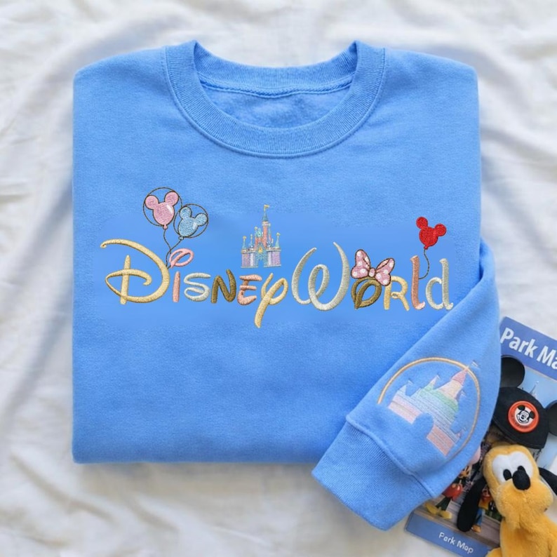 Puede incluir: Sudadera azul claro con el texto bordado "Disney World" en dorado. El dise&ntilde;o incluye globos de Mickey Mouse, un castillo y un lazo de Minnie Mouse. El pu&ntilde;o de la manga presenta un dise&ntilde;o de castillo.