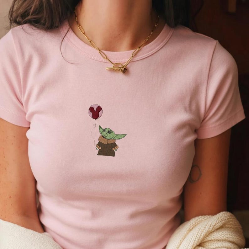 Puede incluir: Camiseta rosa claro con una imagen bordada de un personaje verde parecido a un extraterrestre sosteniendo un globo rojo. El personaje lleva un abrigo marr&oacute;n. El globo tiene forma de cabeza de Mickey Mouse.