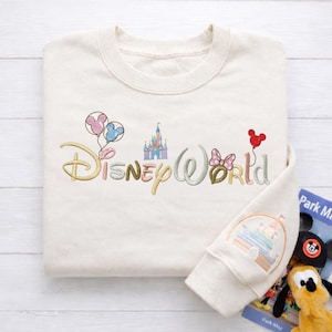 Puede incluir: Sudadera de color crema bordada con "Disney World" en colores pastel. El dise&ntilde;o incluye globos de Mickey Mouse, un castillo y un lazo de Minnie Mouse. La manga presenta una imagen bordada colorida de un castillo y personajes.