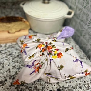 May include: A fabric wrap tied in a knot, featuring a white background with a colorful floral pattern of orange, yellow, and purple flowers with green leaves. The interior lining of the wrap is a light purple fabric with white polka dots.