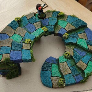 May include: A miniature curved stone staircase model featuring textured blocks painted in shades of blue, teal, and grey. The structure is decorated with small patches of green moss and yellow foliage, with a small figurine standing at the top.