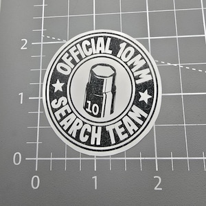 May include: A circular sticker with the text "OFFICIAL 10MM SEARCH TEAM" in a black and white design. The center graphic is a black and white illustration of a socket wrench. The number "10" is visible on the wrench.
