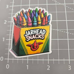 May include: A rectangular sticker featuring a yellow box filled with colorful crayons. The box displays the text "JARHEAD SNACKS" in black letters above a red crayon icon. The sticker is placed on a gray cutting mat with a grid showing a width of 2 inches.