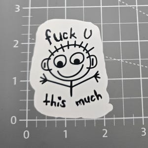May include: A white sticker with a black cartoon figure and the text "fuck U this much". The figure has a smiling face, arms outstretched, and spiky hair. The sticker is on a gray grid.
