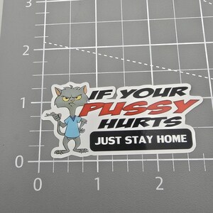 May include: A sticker featuring a cartoon cat with an angry expression, wearing a blue shirt. The sticker has the text "IF YOUR PUSSY HURTS JUST STAY HOME" in black and red.