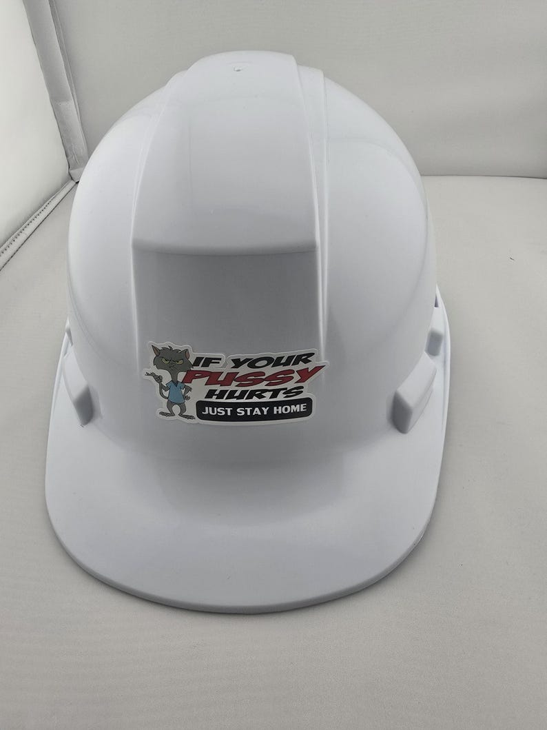 May include: A white hard hat with a sticker that reads "IF YOUR PUSSY HURTS JUST STAY HOME". The sticker features a cartoon cat character. The hard hat is a standard safety helmet.