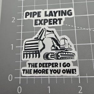 May include: A white sticker with black text and a black and white excavator illustration. The text reads "PIPE LAYING EXPERT" and "THE DEEPER I GO THE MORE YOU OWE!"