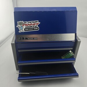 May include: A blue metal toolbox with two open drawers. The toolbox has a silver stripe and the text "P.I.T. Master Box". A sticker on the top reads "IF YOUR PUSSY HURTS JUST STAY HOME". A level and screwdriver are visible in the drawers.
