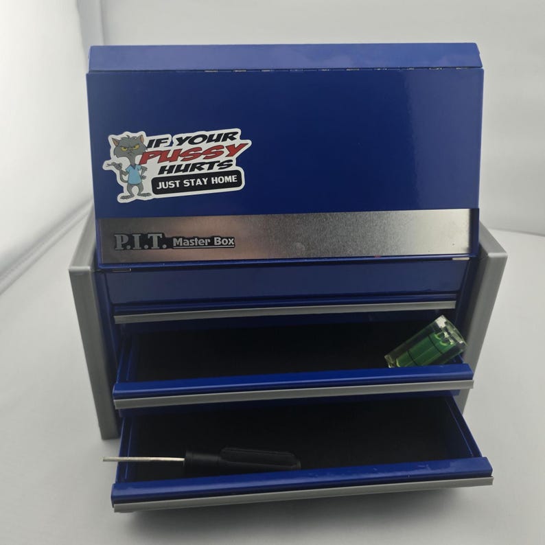 May include: A blue metal toolbox with two open drawers. The toolbox has a silver stripe and the text "P.I.T. Master Box". A sticker on the top reads "IF YOUR PUSSY HURTS JUST STAY HOME". A level and screwdriver are visible in the drawers.