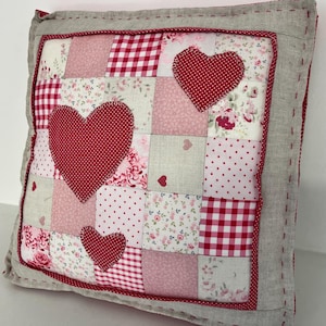 May include: A patchwork cushion with a red and white gingham pattern, pink floral fabric, and three red hearts. The cushion is surrounded by a light grey border.