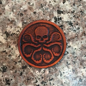 May include: A round, brown leather patch featuring a skull with tentacles, encircled by a border. The design is embossed, creating a raised effect. The patch is likely a decorative item or emblem.