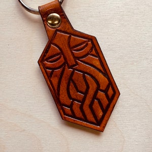 Könnte beinhalten: Ein brauner Leder-Schlüsselanhänger mit einem geschnitzten Design eines Gesichts mit Bart. Der Schlüsselanhänger hat einen goldenen Metallring.